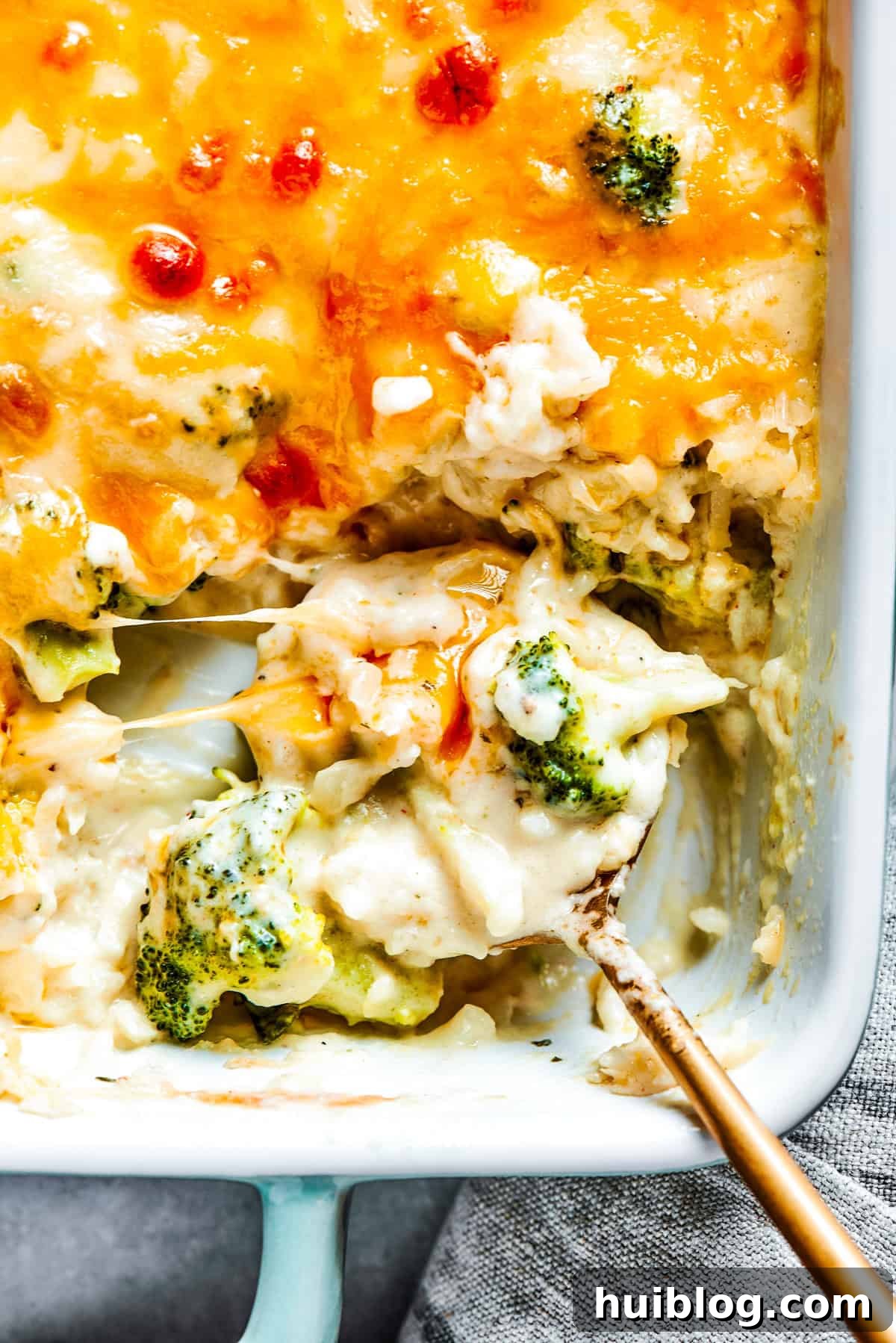 From-scratch Broccoli & Rice Casserole folds tender broccoli and fluffy white rice into a gorgeously creamy three-cheese sauce. A serving spoon lifting a generous portion of cheesy broccoli rice casserole from a hot baking dish, showing off the melted cheese and tender ingredients.