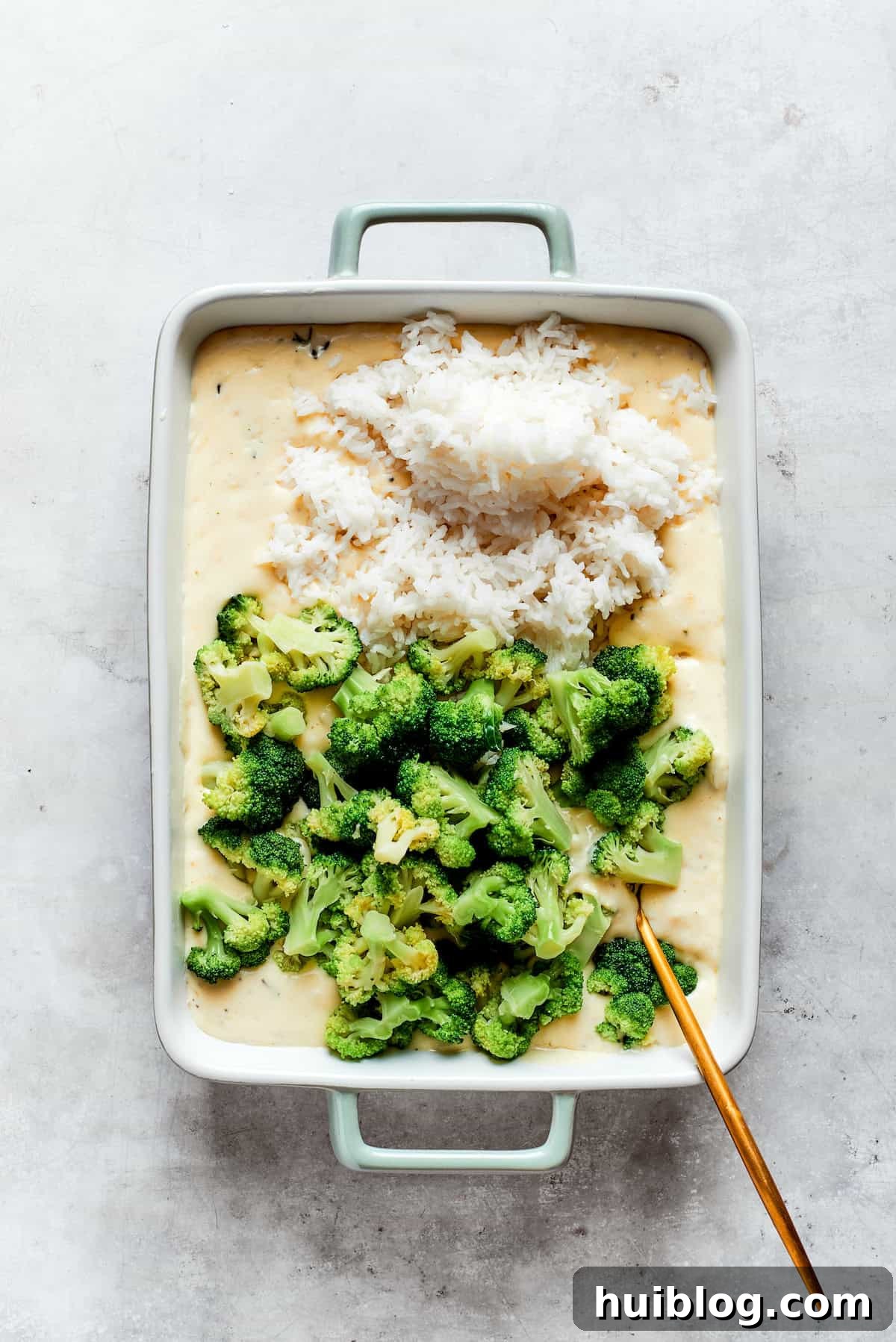 From-scratch Broccoli & Rice Casserole folds tender broccoli and fluffy white rice into a gorgeously creamy three-cheese sauce. A baking pan holds the rich cheese sauce, waiting for the addition of cooked broccoli and rice before being gently combined.