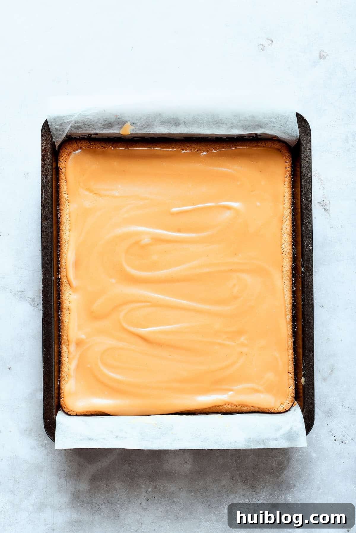 The freshly cooked caramel sauce is being poured evenly over the cooled shortbread crust in a baking pan.