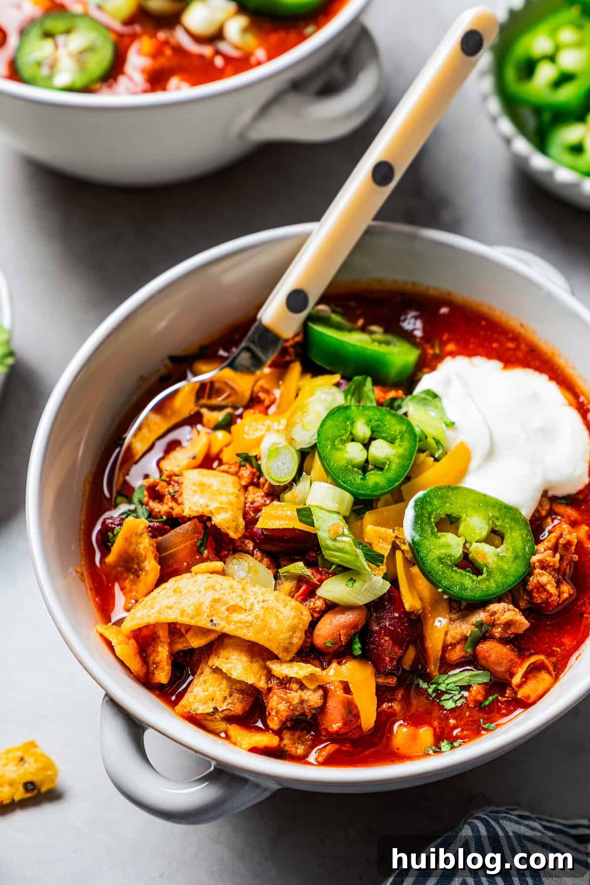 A bowl of turkey chili garnished with sour cream, jalapeños, and corn chips, with a second bowl in the background.