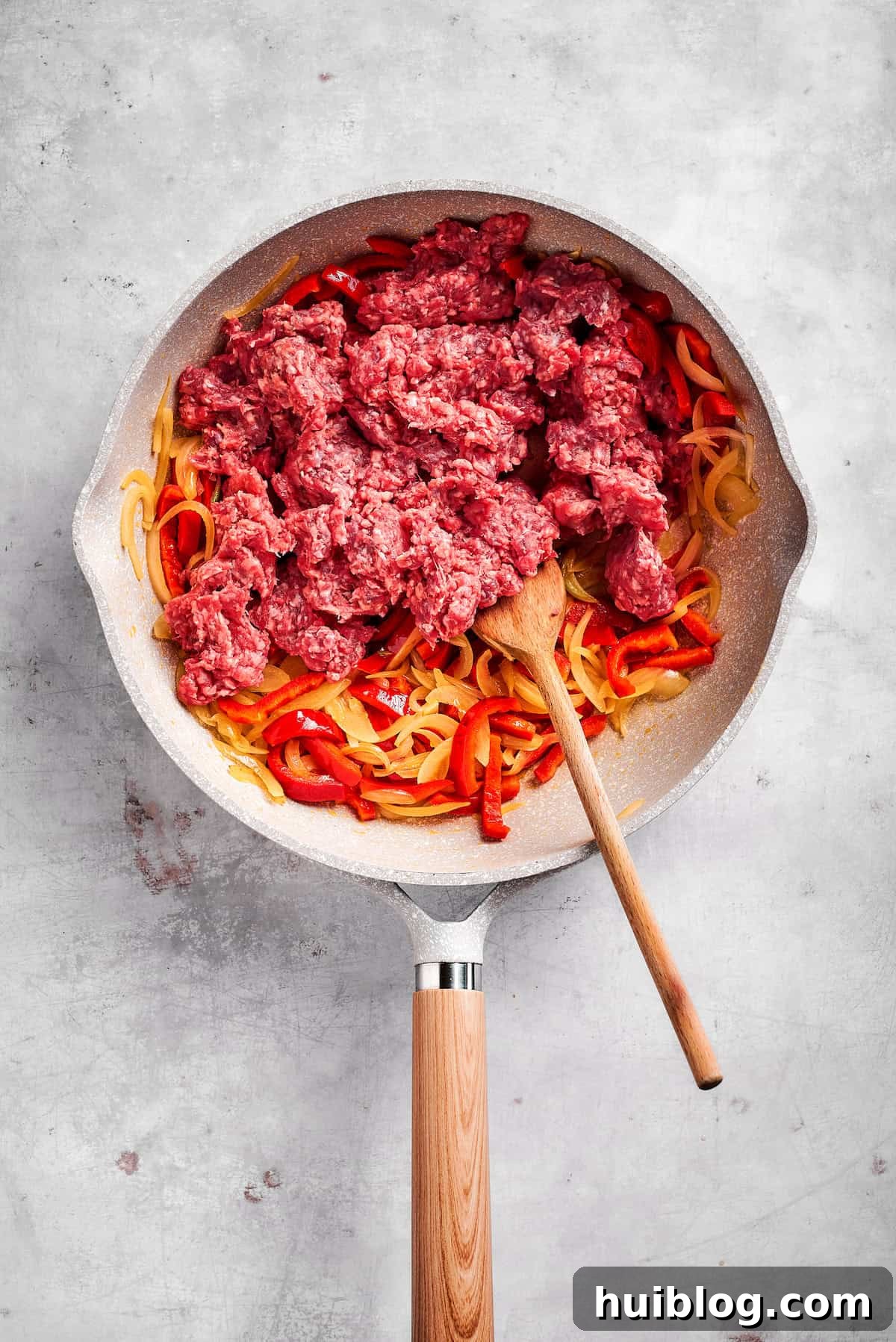 Adding ground beef to onions and bell peppers sautéing in a skillet.