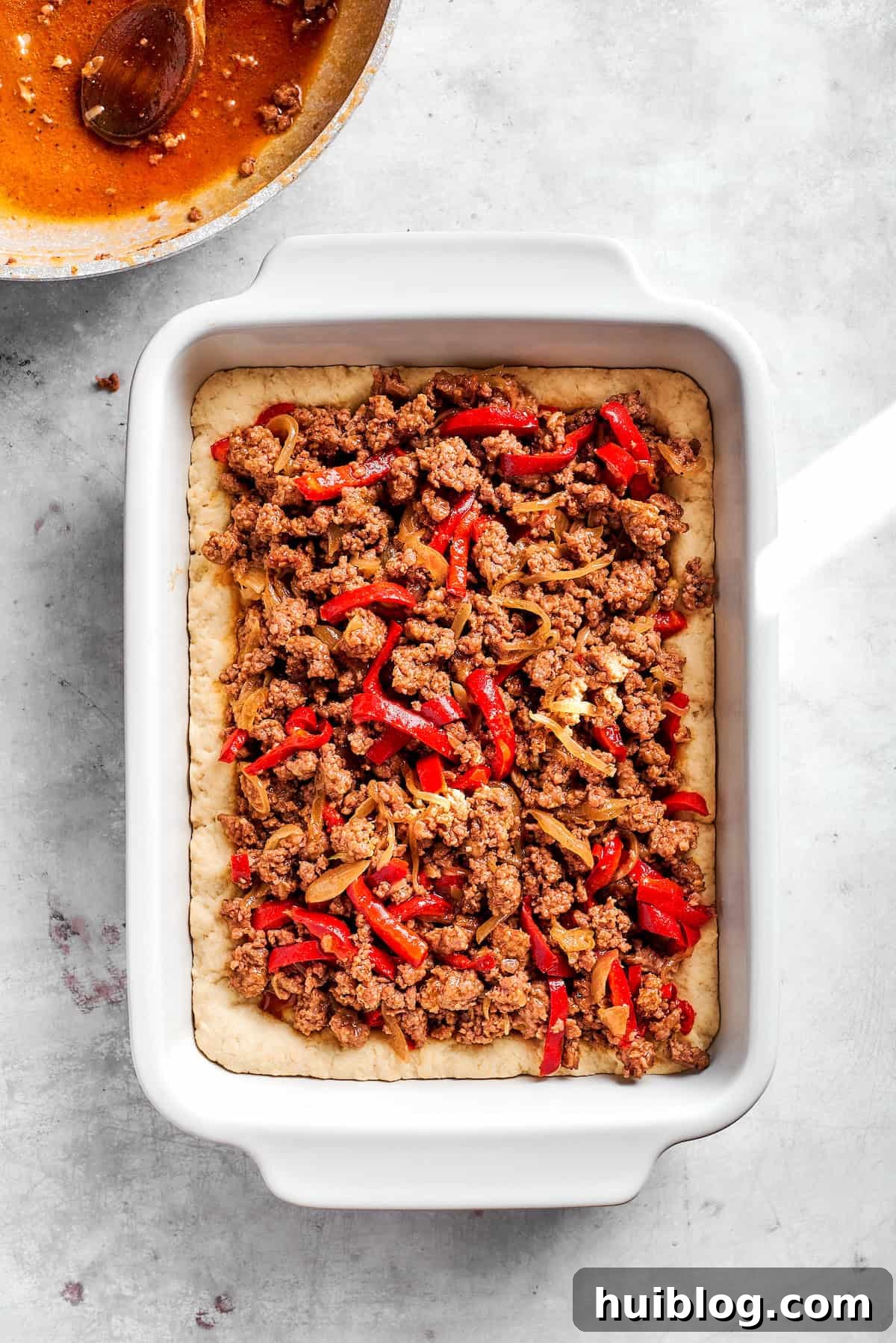 Adding the cooked ground beef filling over the biscuit dough in the baking dish.