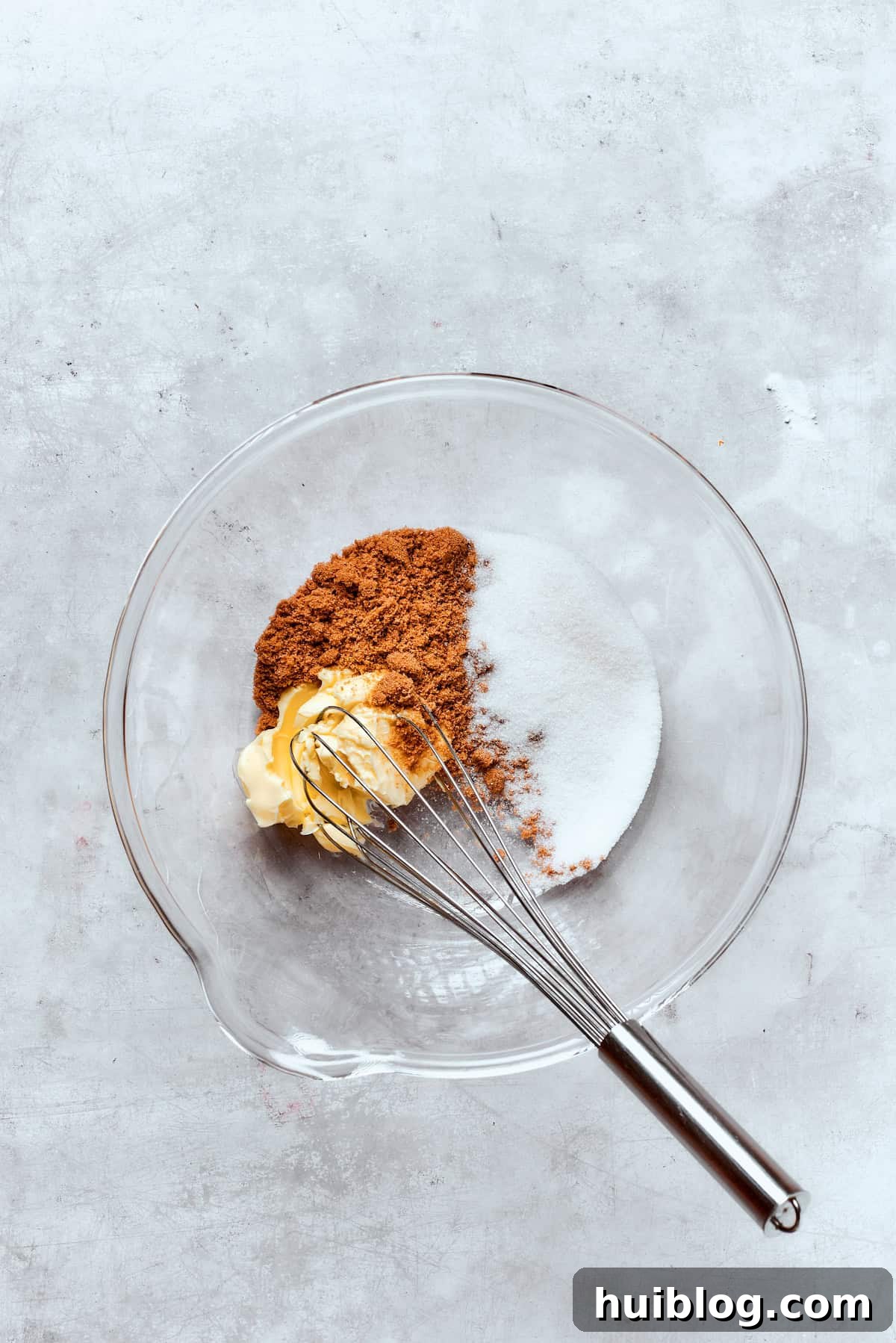 A whisk combines softened butter with granulated and brown sugar in a mixing bowl until light and fluffy.