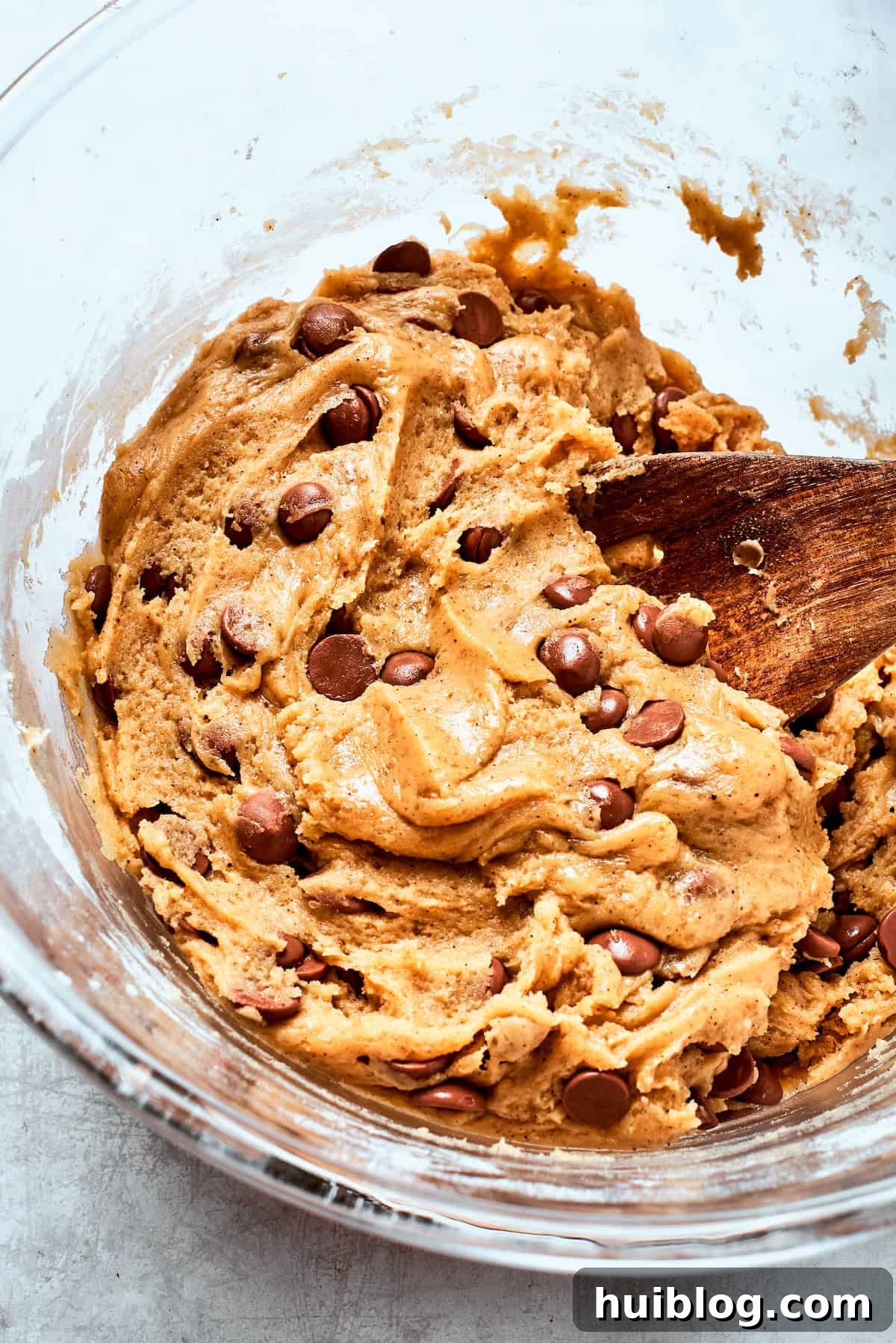 A wooden spoon carefully folds an abundance of semi-sweet chocolate chips into the rich brown butter cookie dough.