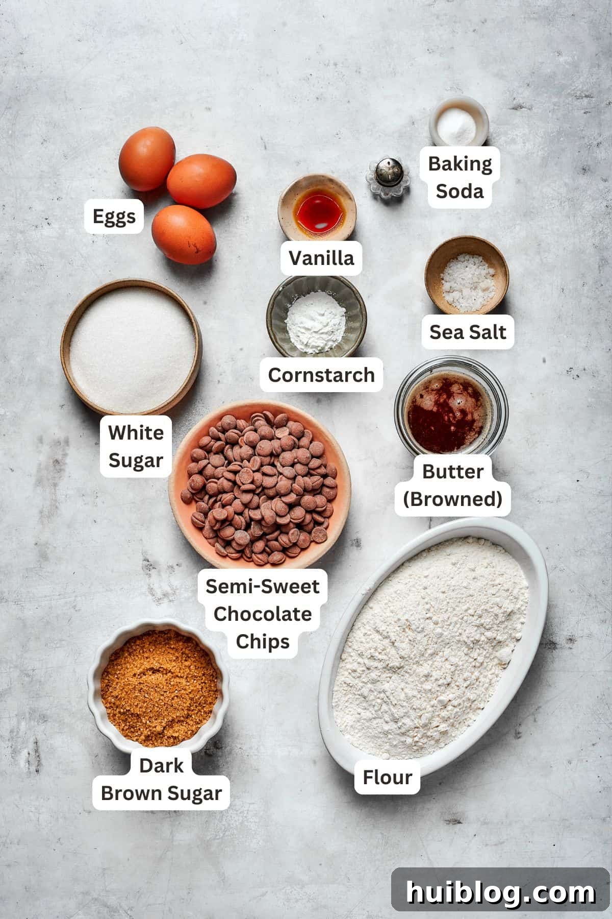 Labeled ingredients for brown butter chocolate chip cookies, including butter, dark brown sugar, and chocolate chips, arranged artfully on a surface.