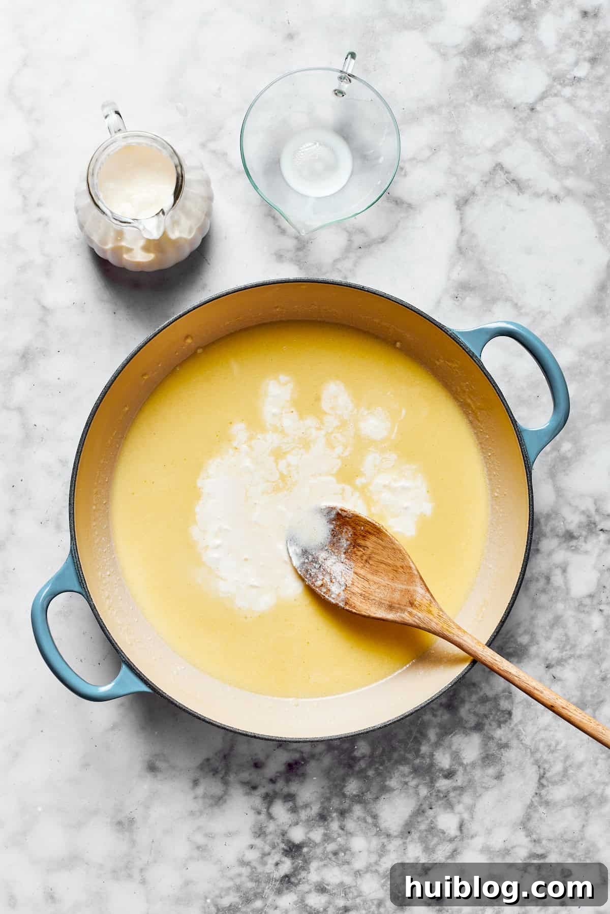 Add milk & cream. A saucepan containing a roux with milk and heavy cream being gradually whisked in, forming the creamy base of the garlic parmesan sauce.