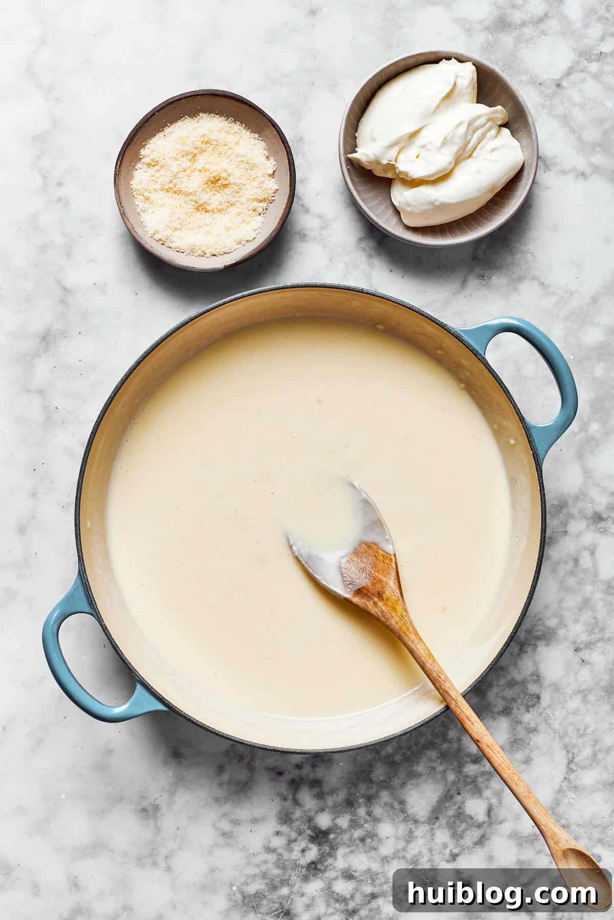 Whisk until smooth. A creamy, thickened sauce in a saucepan, next to bowls of shredded Parmesan cheese and cubed cream cheese, ready to be incorporated.