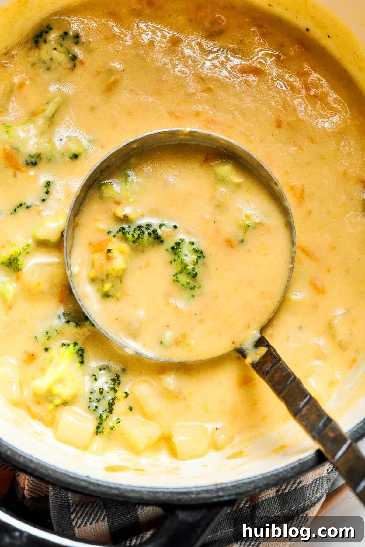 A ladle scooping a generous portion of creamy broccoli cheese soup from a large cooking pot, showcasing its rich texture and embedded vegetables.
