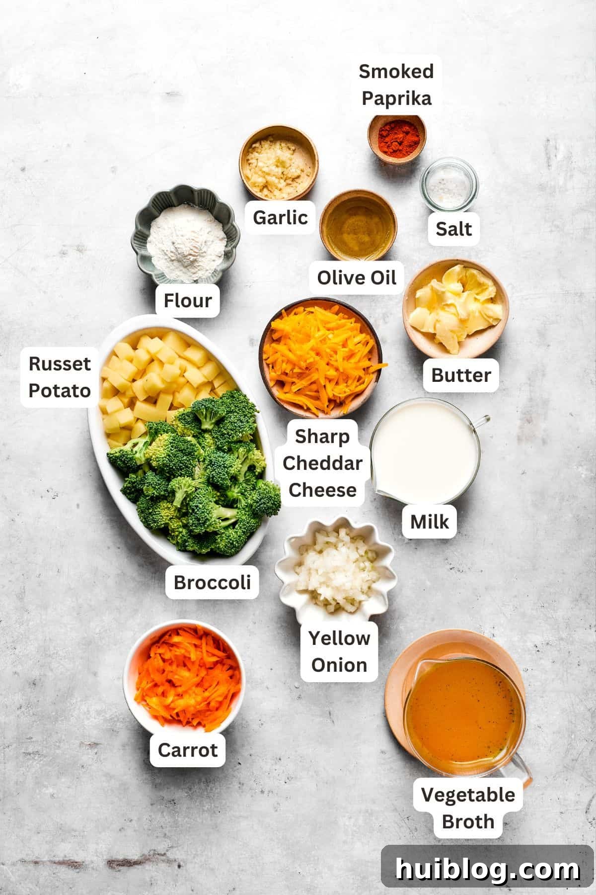 A flat lay of fresh ingredients for broccoli cheese soup, including broccoli florets, carrots, onions, potatoes, garlic, milk, broth, cheese, and seasonings.