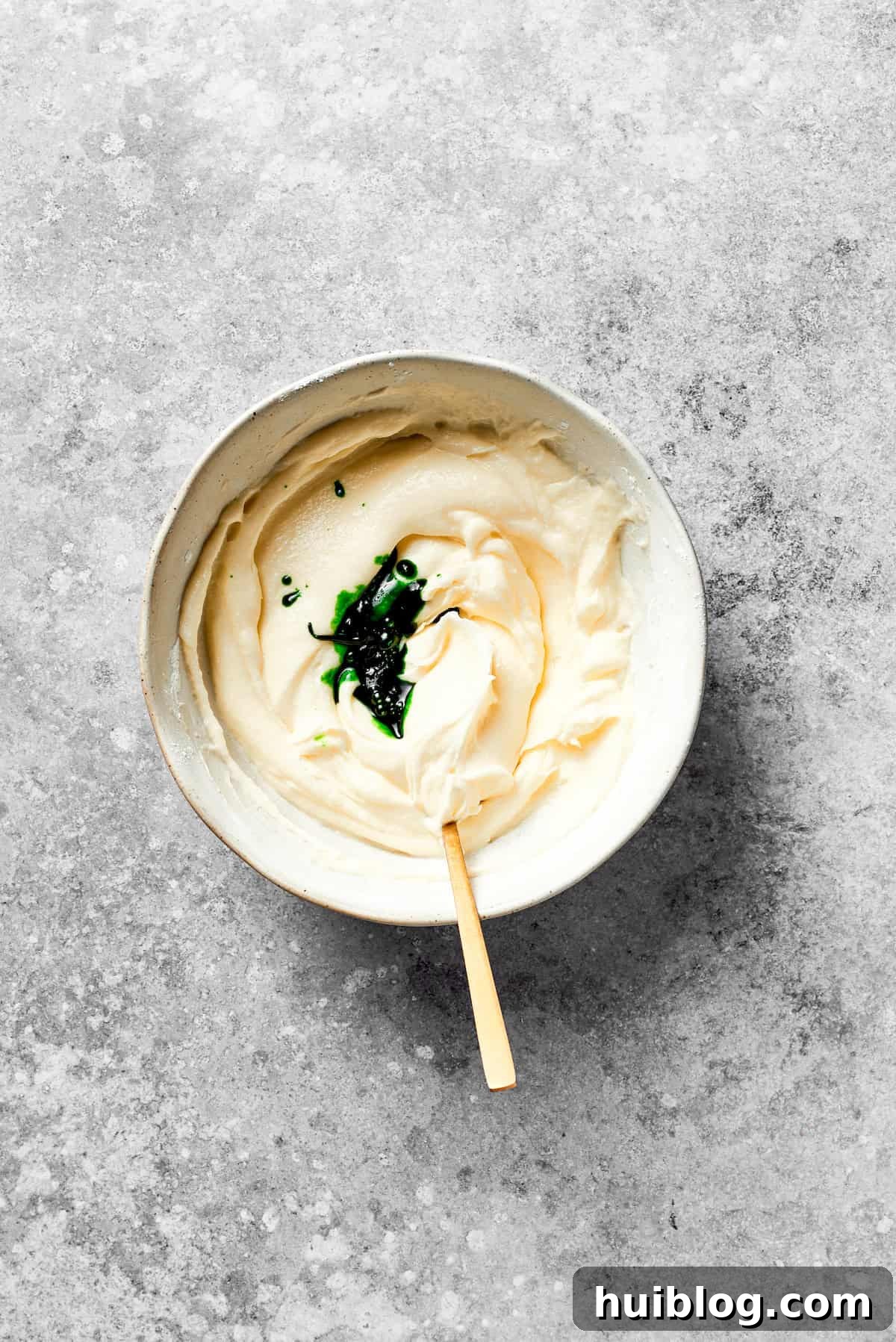 Green food coloring is added to a bowl of white cream cheese icing.
