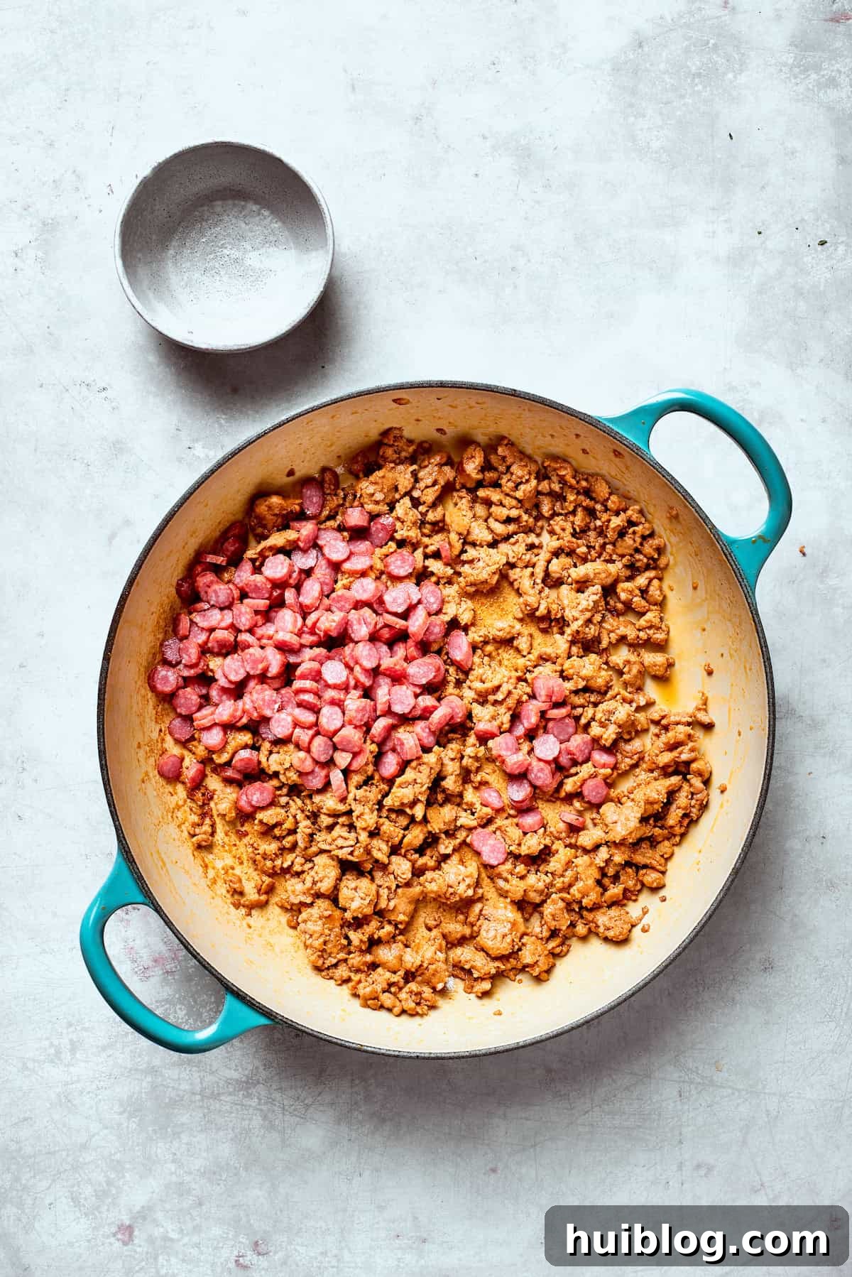 Pepperoni slices are scattered over browned Italian sausage in a large skillet, ready to be stirred.
