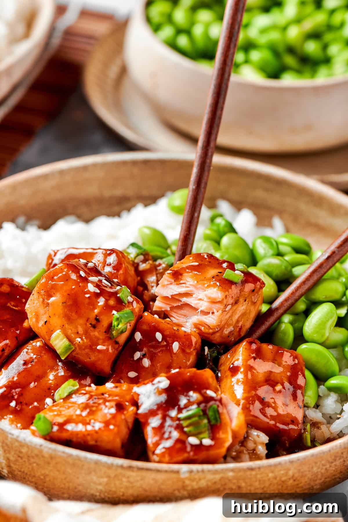 A pair of chopsticks gently picks up a piece of glistening teriyaki salmon from a bowl, highlighting its perfect glaze and tender texture.
