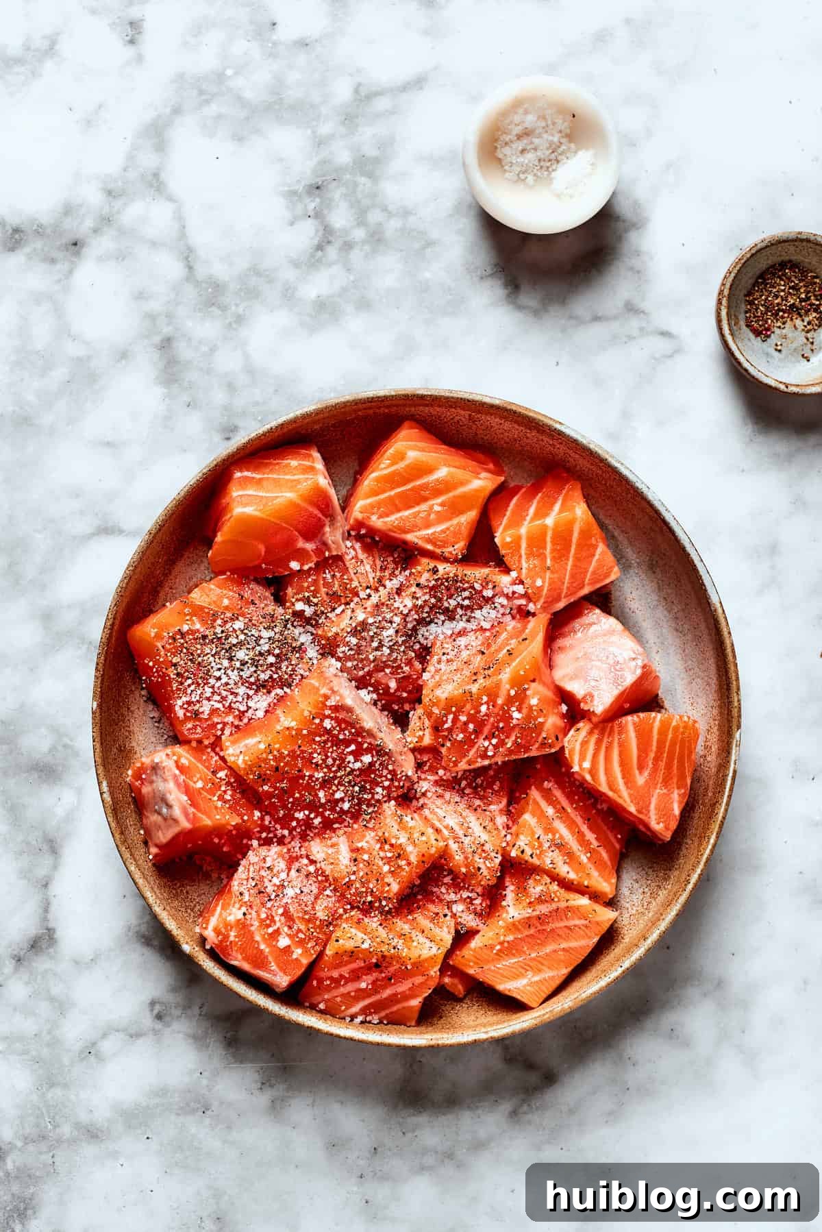 Cubed salmon pieces are seasoned generously with salt and pepper on a cutting board.