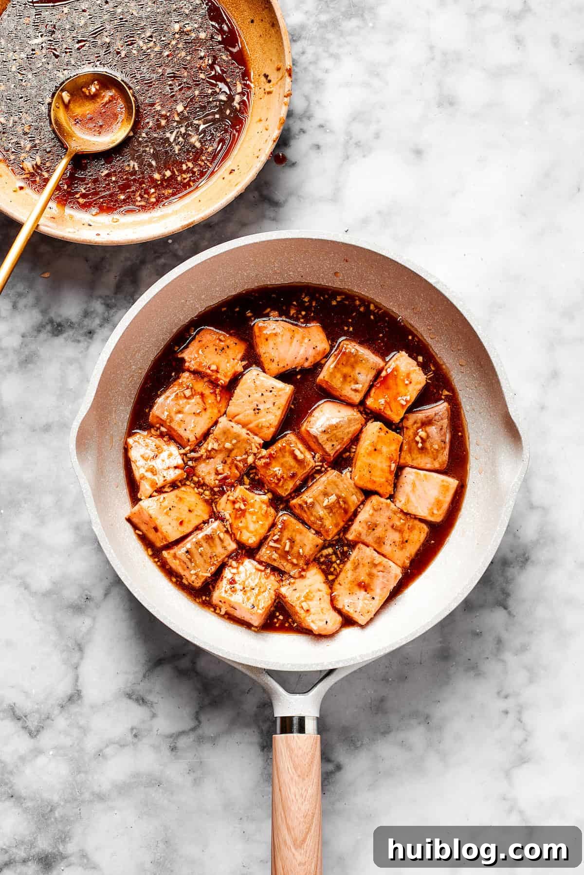 The thickened teriyaki sauce is poured over the seared salmon in a skillet, coating each piece.