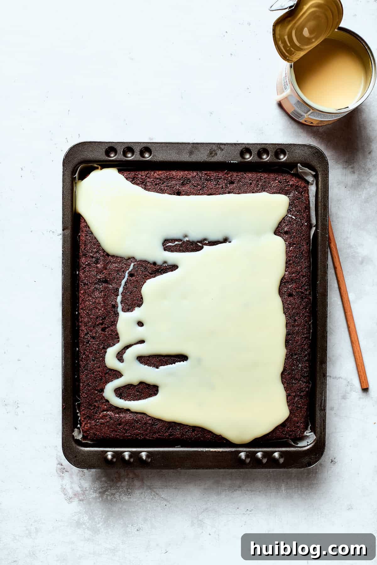 Sweetened condensed milk being poured slowly over the poked surface of a chocolate cake.