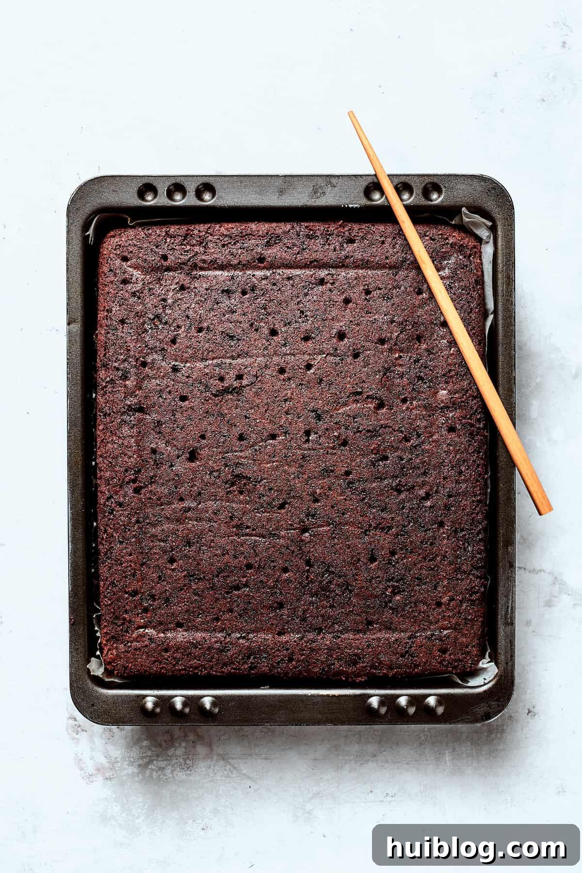Freshly baked chocolate cake cooling in a tin, with poked holes on its surface.