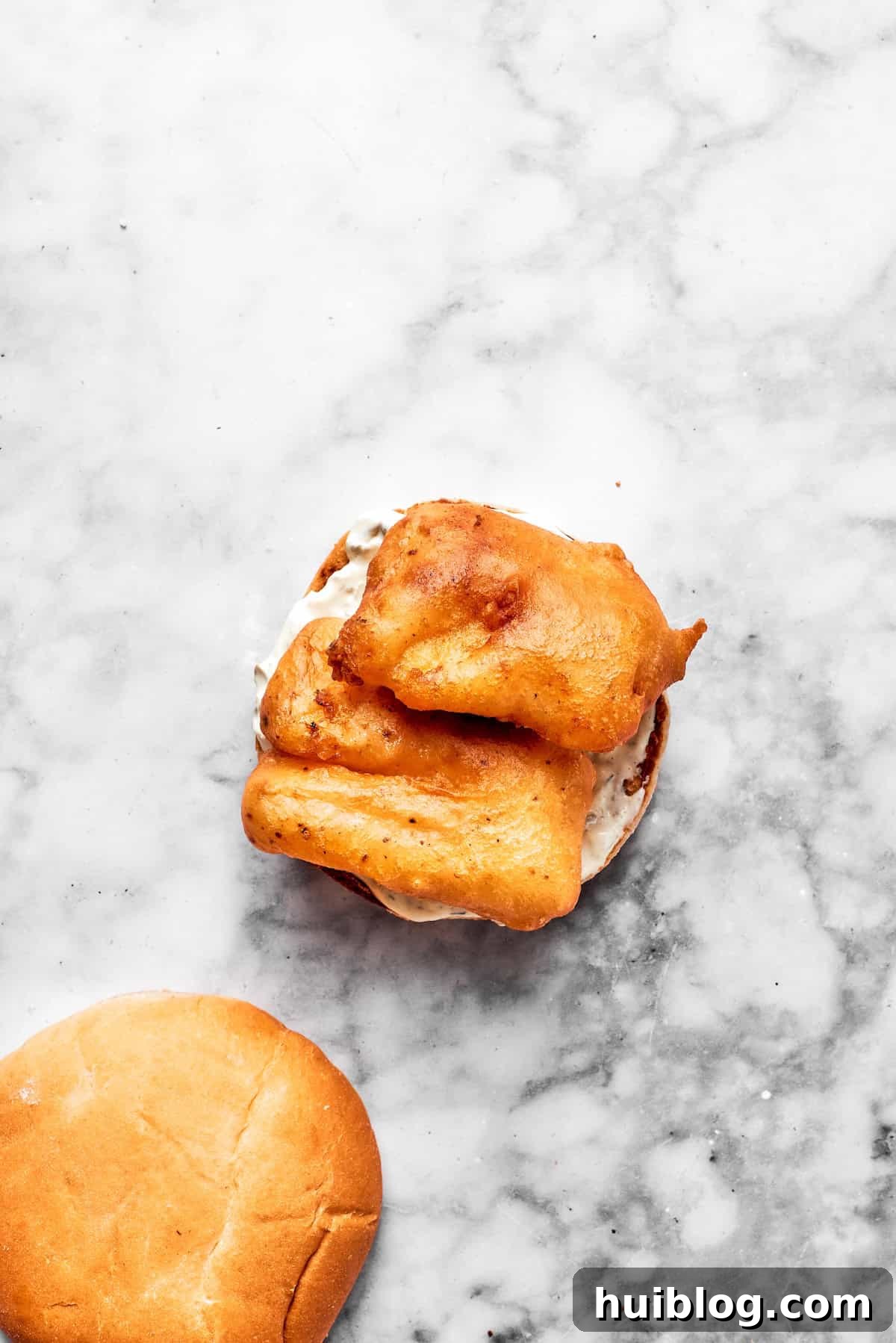 This crispy, crunchy fish sandwich is served on toasty sweet rolls with lemony tartar sauce and a squeeze of lime! It's quick and easy to make! A freshly fried, golden-crisp fish fillet is carefully placed on the sauced bottom half of a toasted brioche bun, forming the base of the sandwich.