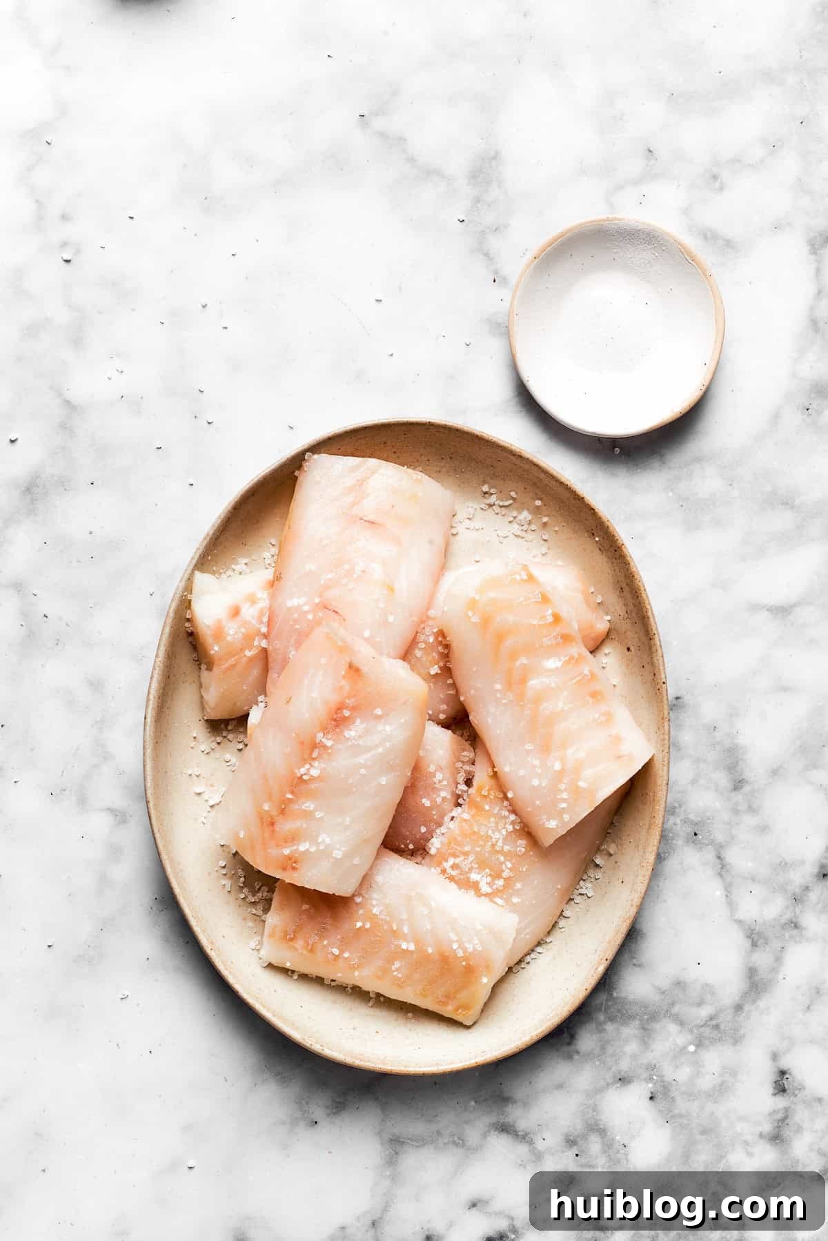 This crispy, crunchy fish sandwich is served on toasty sweet rolls with lemony tartar sauce and a squeeze of lime! It's quick and easy to make! Pieces of fresh white fish fillets are laid out on a plate, lightly seasoned with salt, ready for the next step in preparing the crispy fish sandwich.