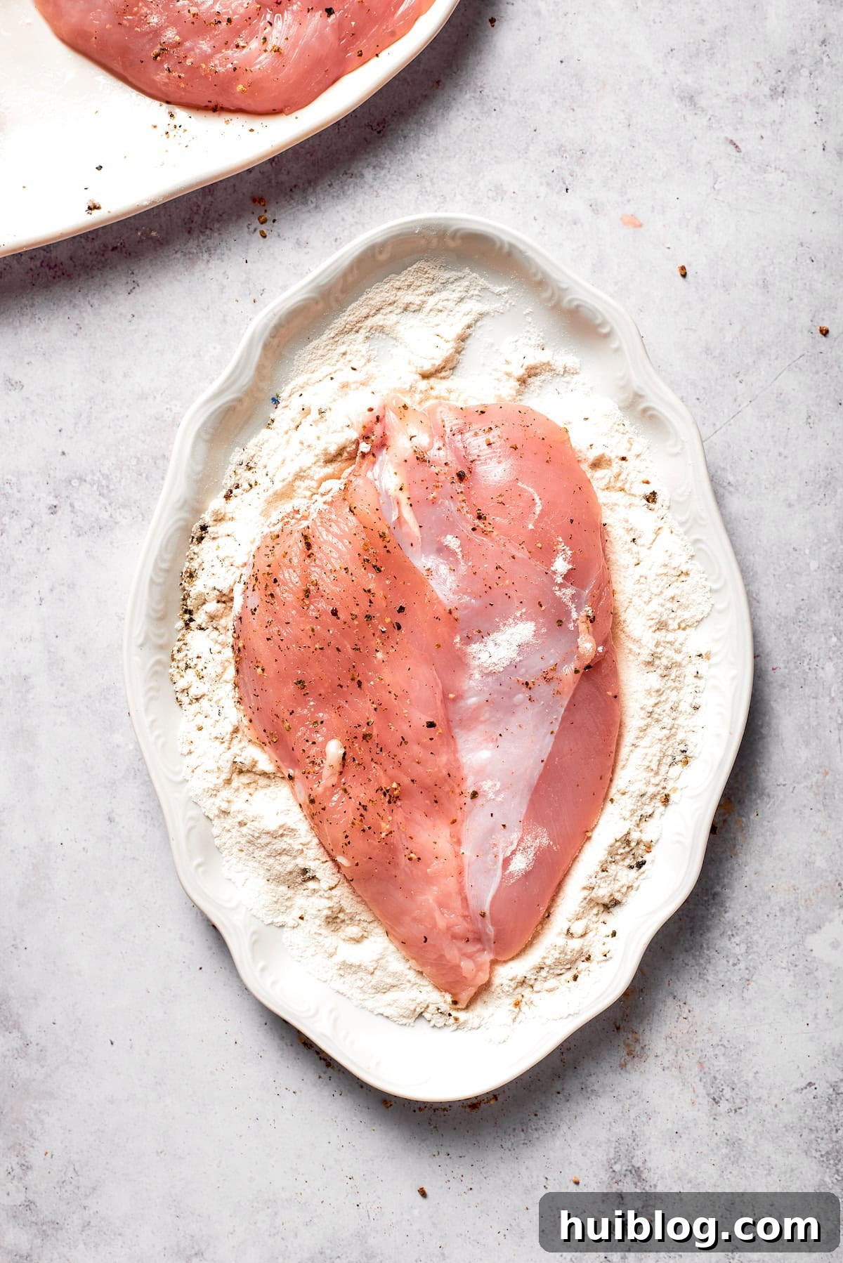A seasoned chicken cutlet being pressed into the flour mixture, ensuring an even and light coating.