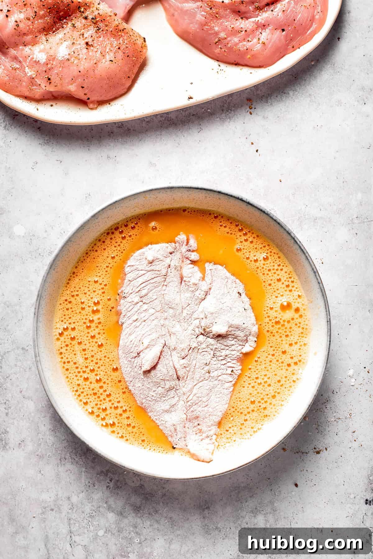 A floured chicken cutlet being submerged into the whisked egg wash, ensuring full coverage.