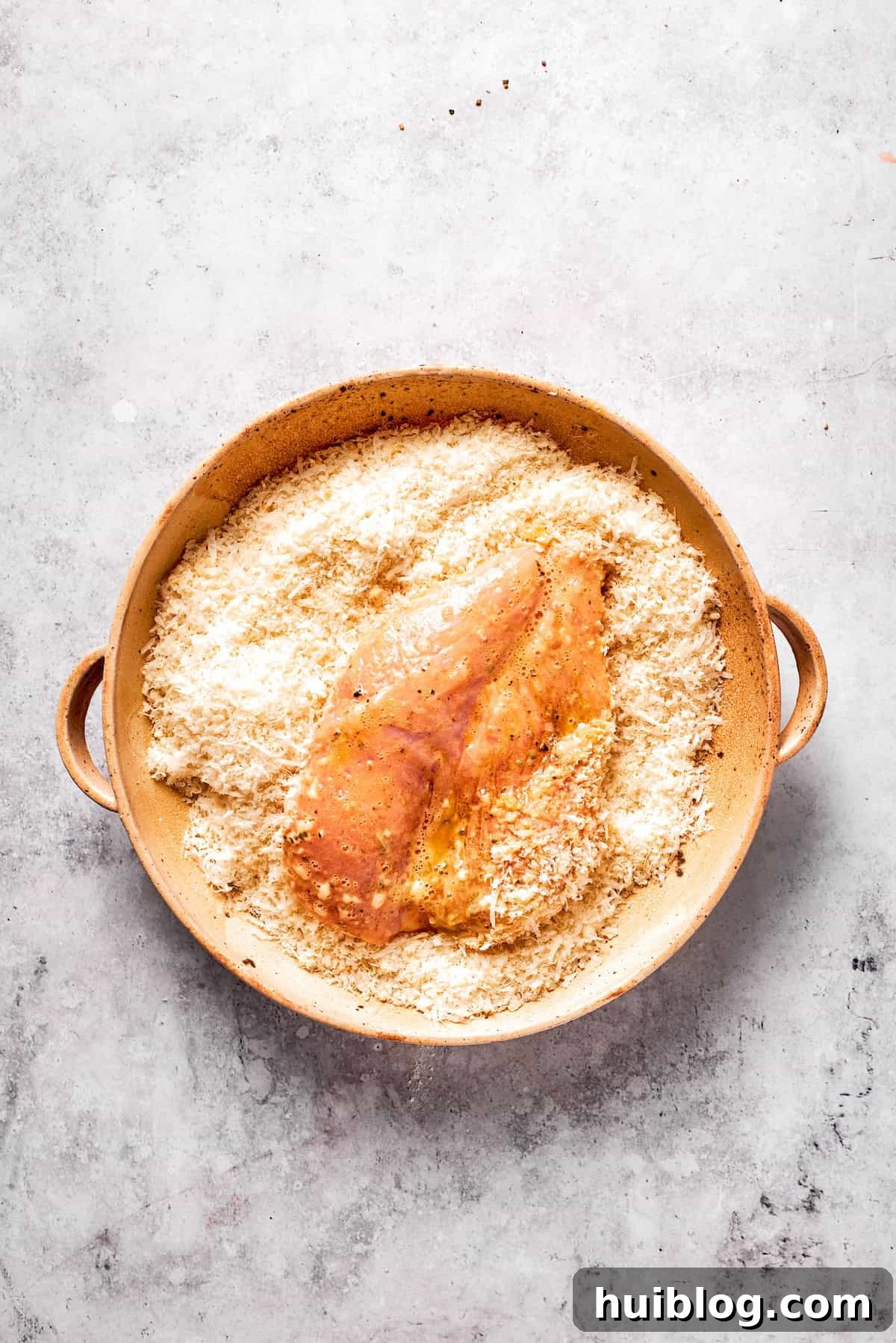 An egg-coated chicken cutlet being placed into the breadcrumb and cheese mixture, ready to be firmly pressed.