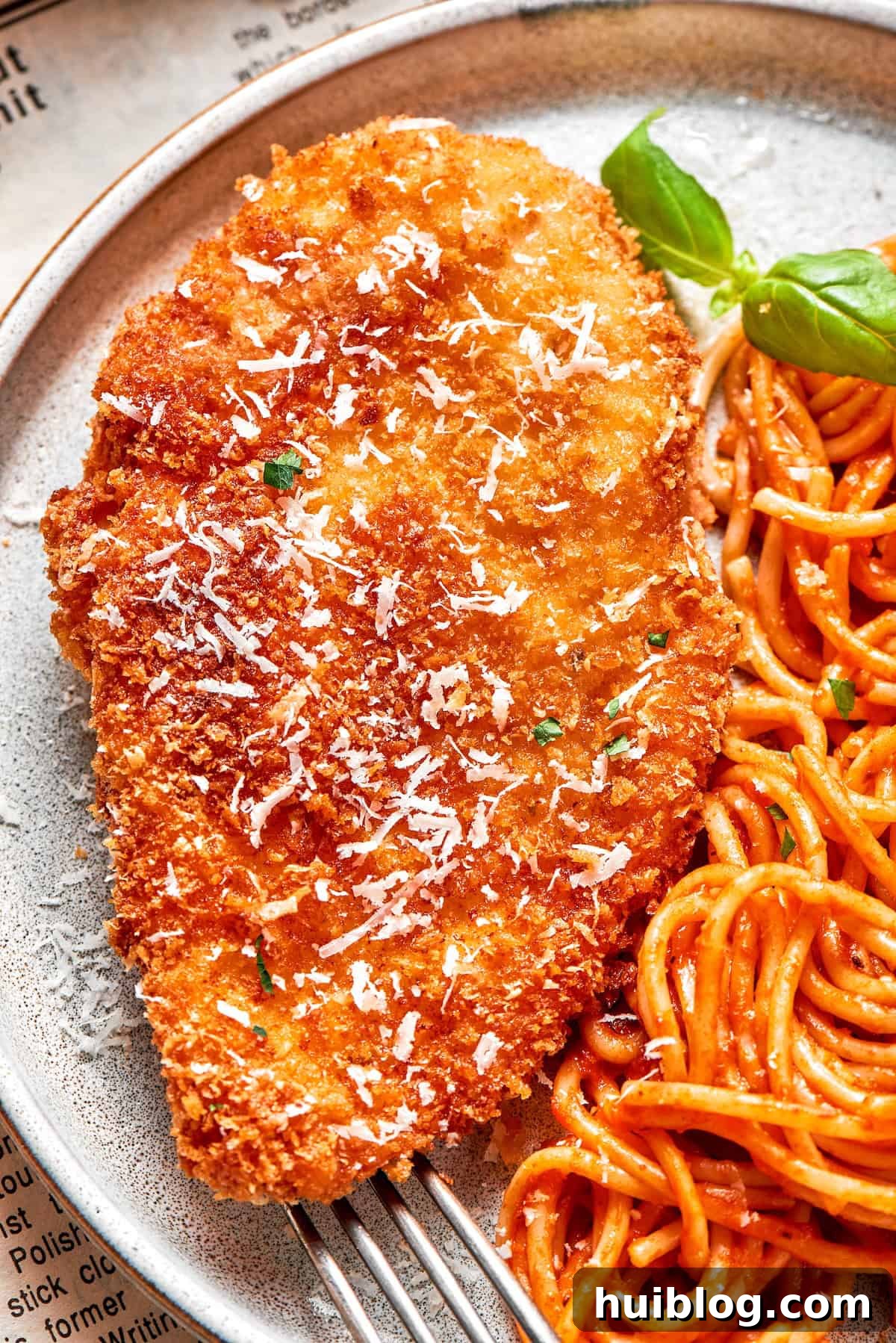 A close-up shot of a large, perfectly golden-brown Crusted Chicken Romano cutlet, showcasing its texture, resting on a bed of savory spaghetti.