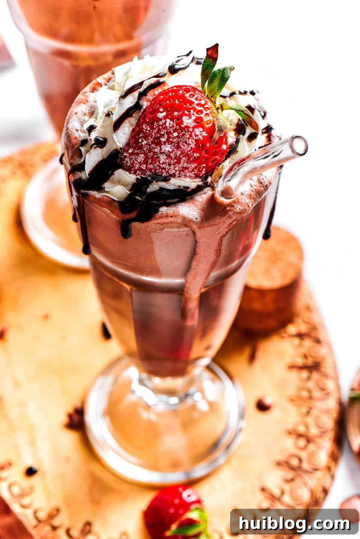 A perfectly prepared chocolate milkshake served in a tall, elegant glass and garnished with a generous dollop of whipped cream, a vibrant fresh strawberry, and a delicate drizzle of chocolate sauce.
