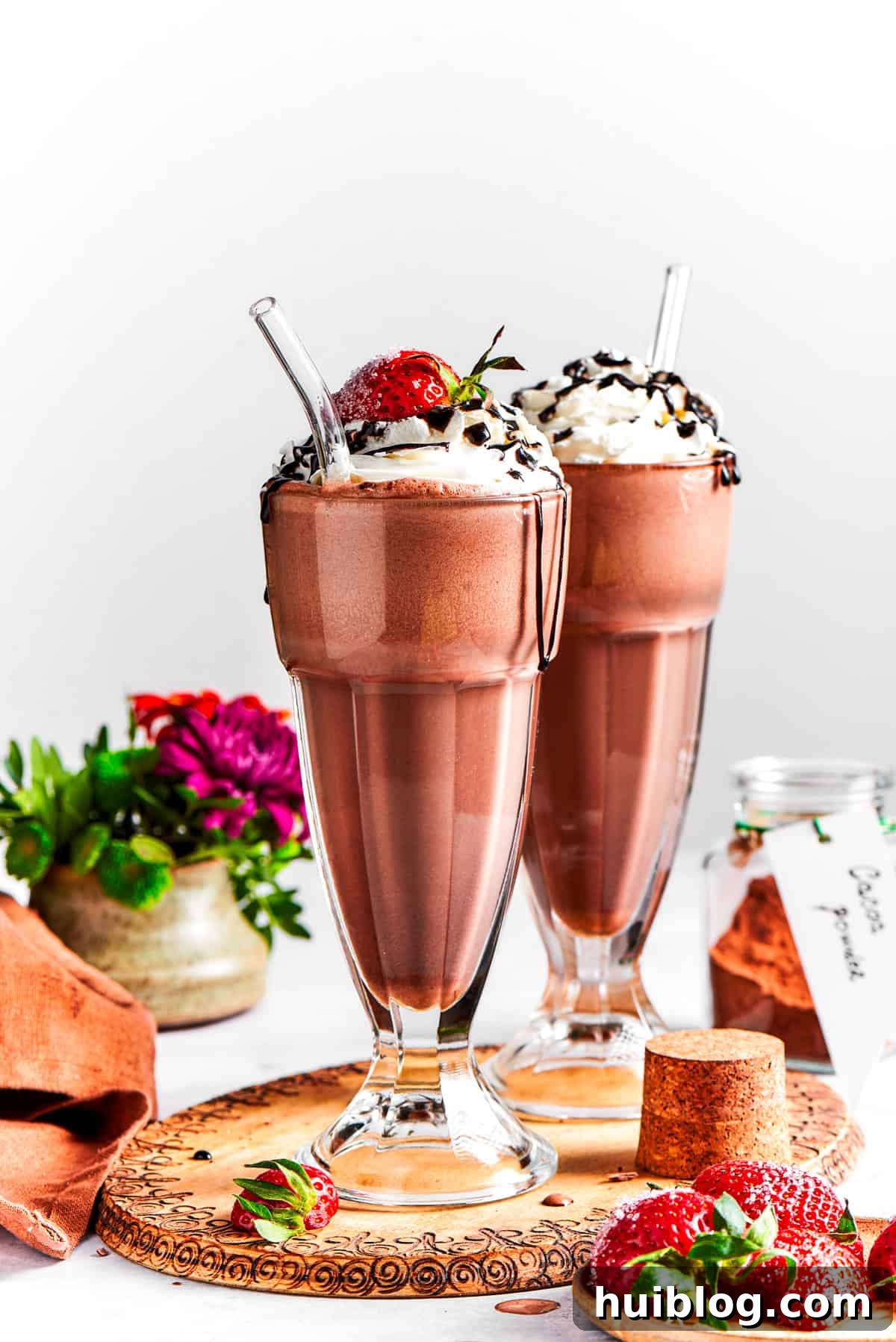 Two tall glasses of rich chocolate milkshake, garnished with whipped cream, fresh strawberries, and chocolate drizzle, sit invitingly on a rustic wooden tray, ready to be enjoyed.