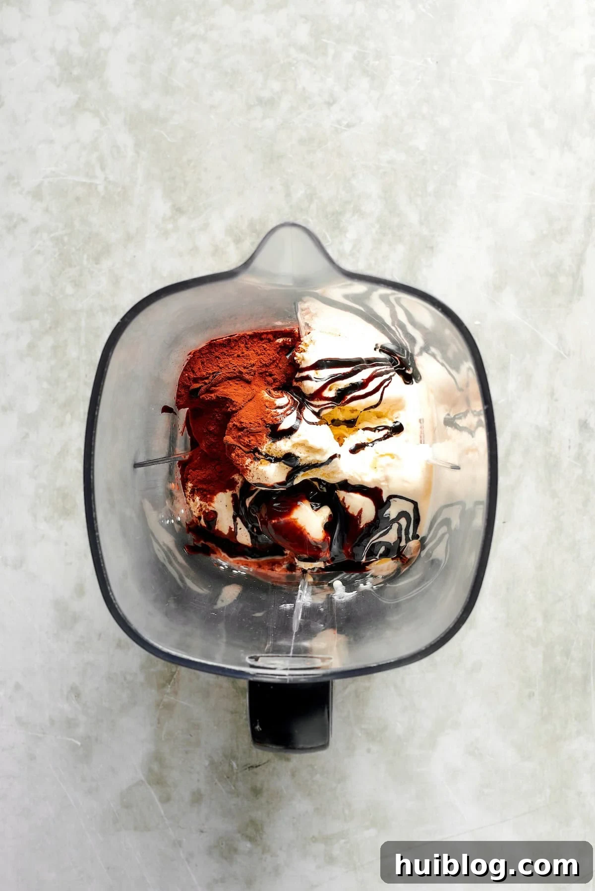 Ingredients for chocolate milkshakes are added to a blender. Heavy cream is poured in, followed by scoops of ice cream, then cocoa powder and chocolate sauce, layered for optimal blending.