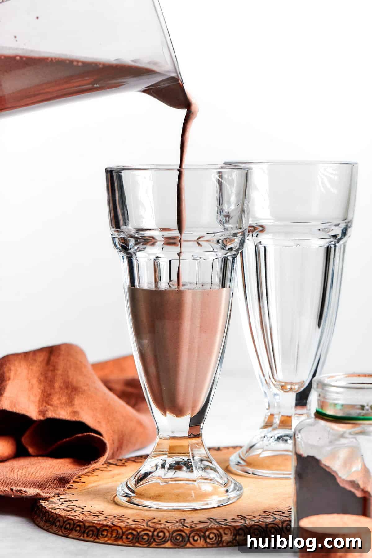A stream of thick, rich chocolate milkshake is being poured from a blender into a tall, chilled glass, ready for garnishing.