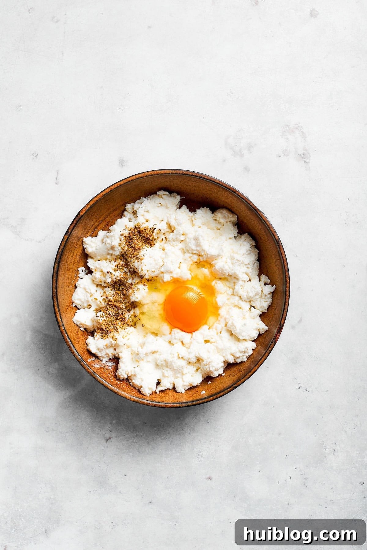 A raw egg is cracked into a bowl of creamy ricotta cheese, ready to be mixed for the lasagna filling.