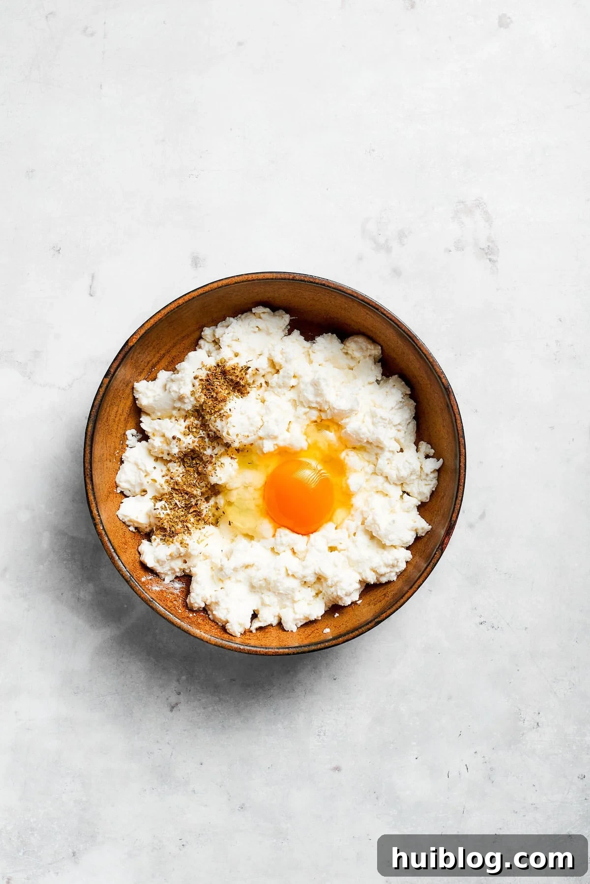 A raw egg being cracked and added to a bowl of ricotta cheese, ready to be mixed for the lasagna filling.