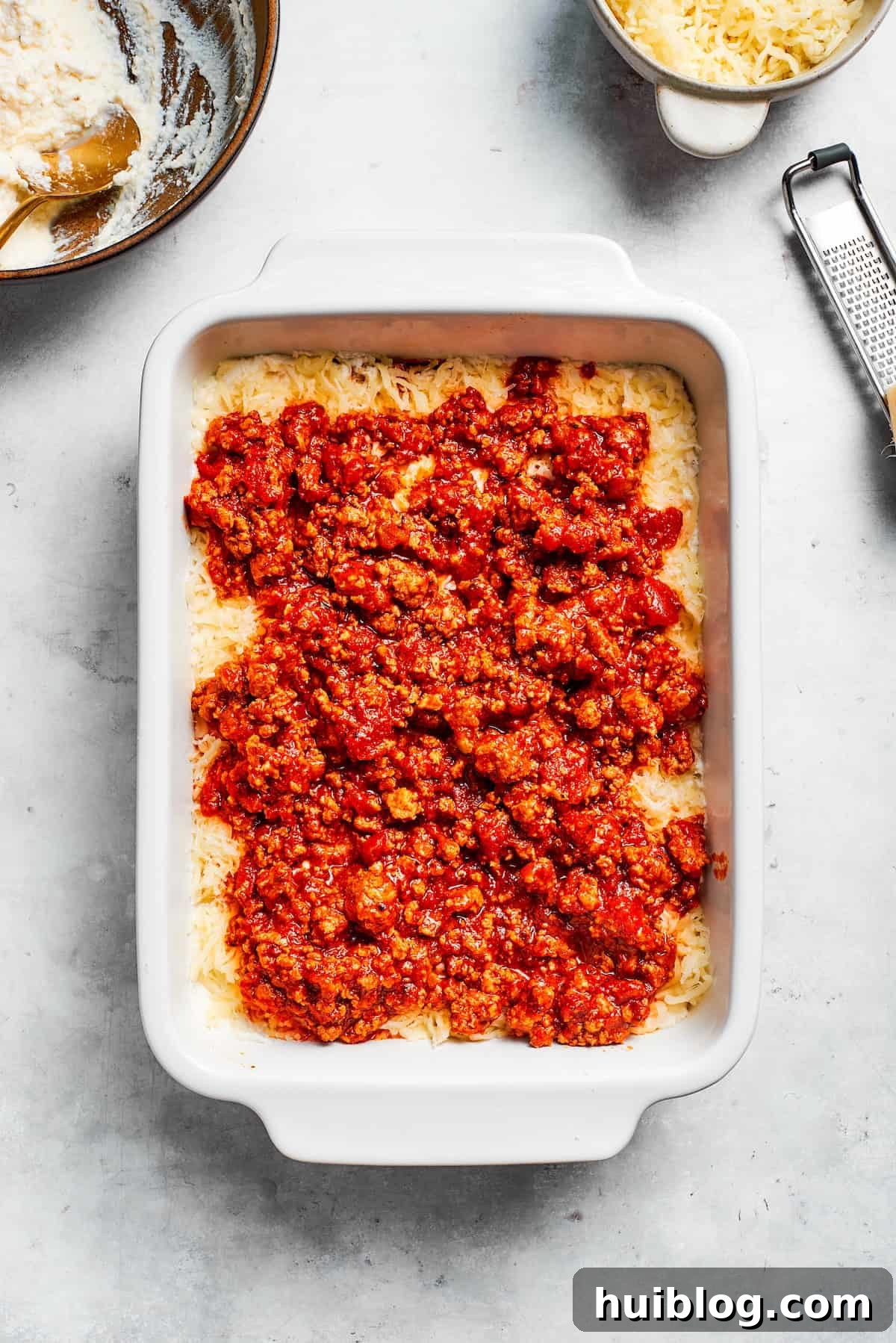 A spoonful of rich meat sauce being spread over a cheesy pasta layer in a partially assembled beef lasagna.