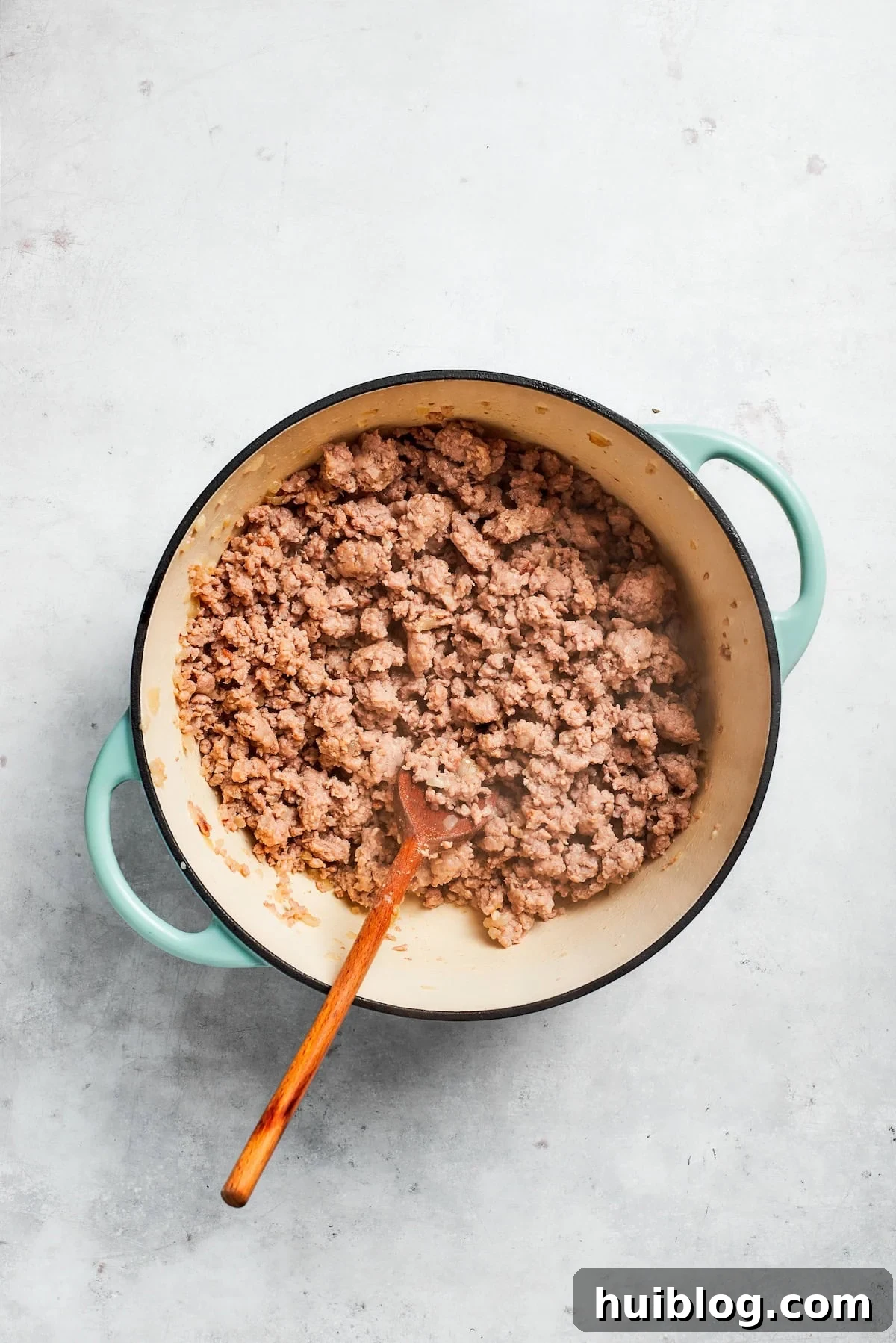 Ground beef browning in a skillet with sautéed onions and garlic, being broken apart with a wooden spoon.