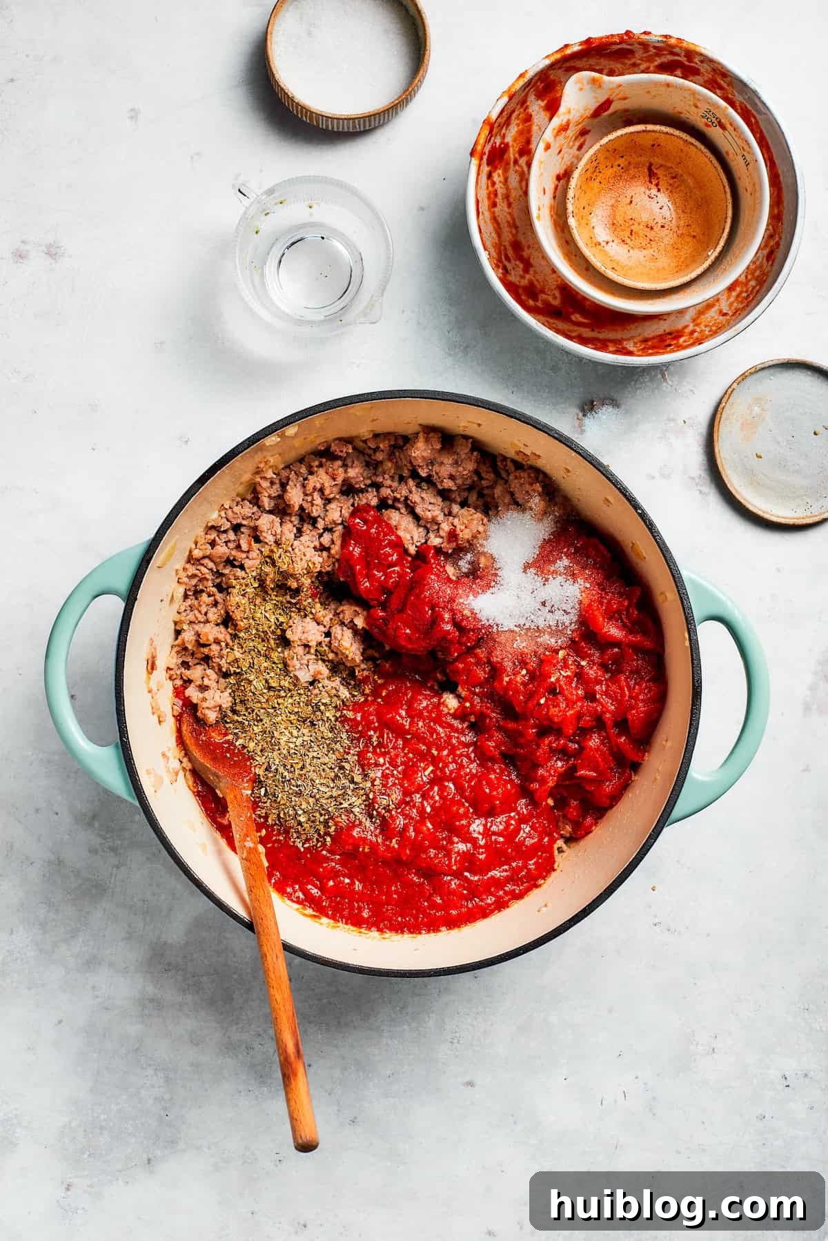 Crushed tomatoes, tomato paste, and various seasonings being added to the browned beef and aromatics in a skillet.