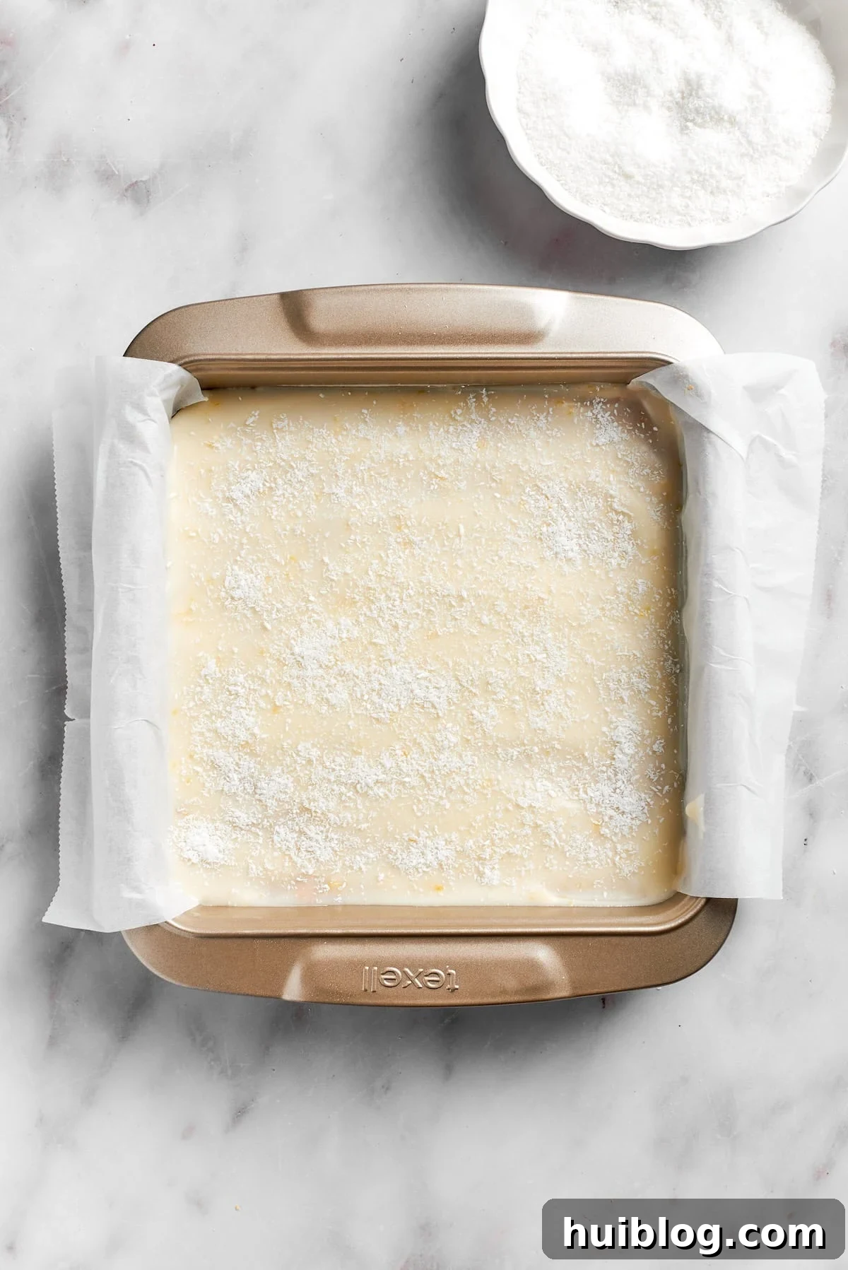 The fully assembled lemon slice, topped with frosting and coconut, resting in a lined pan for chilling.