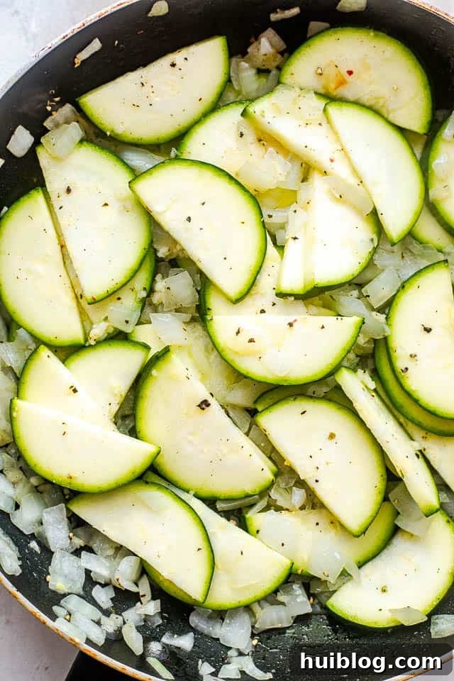 Velvety Zucchini Mushroom Pasta Bake 3 A black skillet on the stovetop, filled with thinly sliced zucchini and diced yellow onion being sautéed until tender.