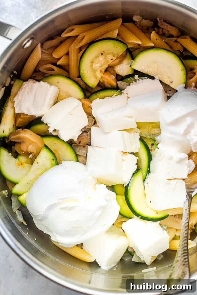 Velvety Zucchini Mushroom Pasta Bake 4 An overhead view of a large silver pot containing cooked penne pasta, sautéed zucchini slices, and cubes of cream cheese, ready to be mixed into the sauce.