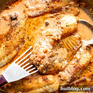 Pan-fried tilapia fillet being lifted from a skillet with a fish spatula, accompanied by creamy tomato sauce.