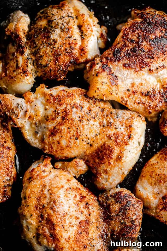 Sticky Apple Cider Chicken Thighs 4 A close-up shot of boneless chicken thighs browning and sizzling in a black skillet on a stovetop, surrounded by herbs.