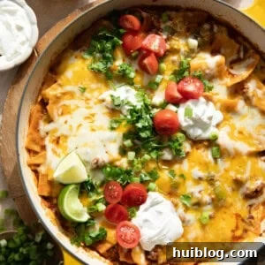 Ground beef enchilada skillet in a braising pan, topped with sliced tomatoes, fresh cilantro, sour cream, and lime wedges.
