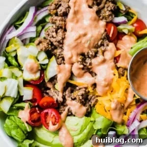 White bowl filled with chopped green lettuce, topped with cooked burger meat, cheese, avocado, tomato, pickle, red onion all drizzled with burger sauce.