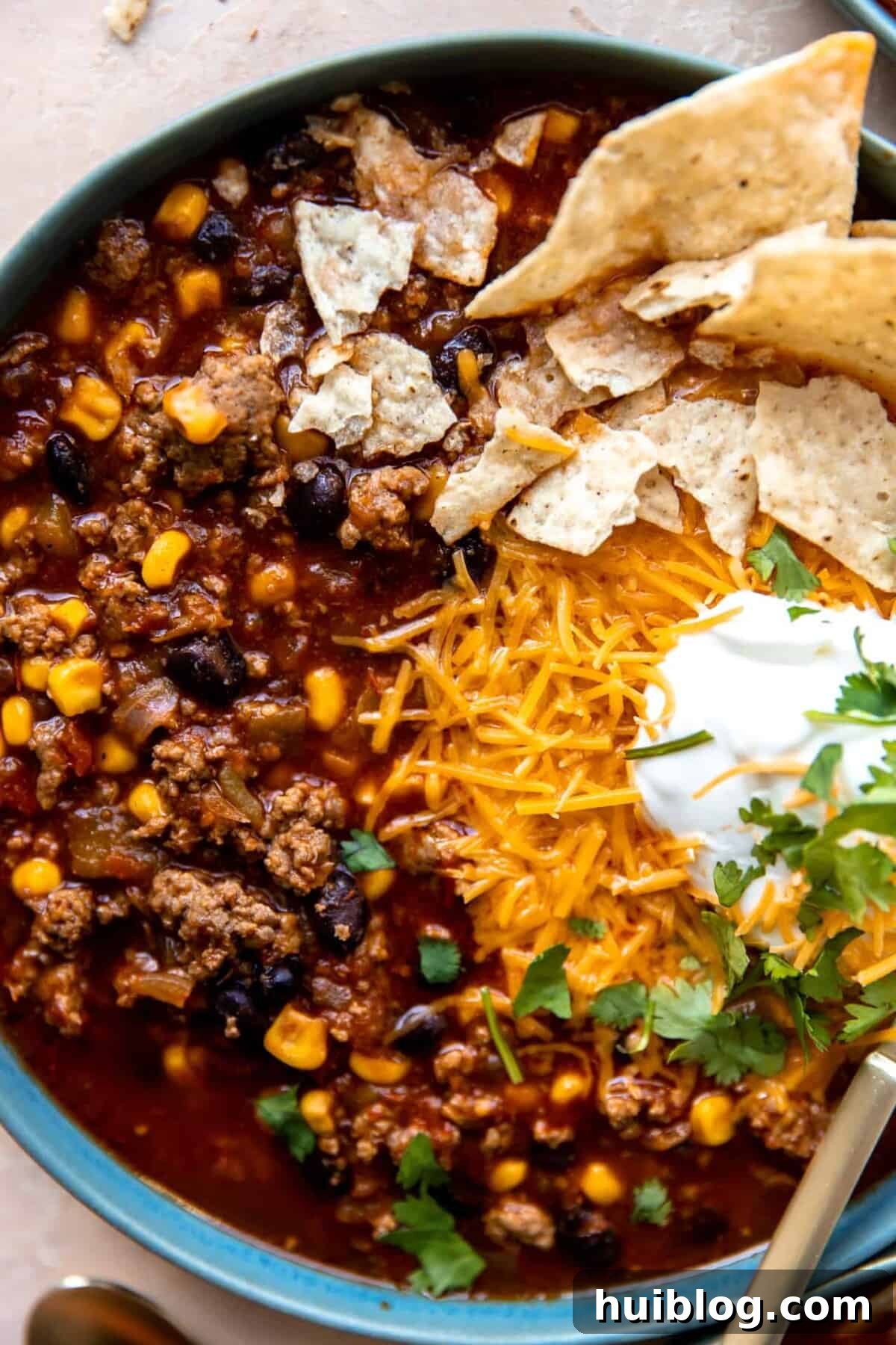 Up close view of a bowl of taco soup showing chunks of beef, beans and corn in a dark red broth topped with melted cheese and sour cream.