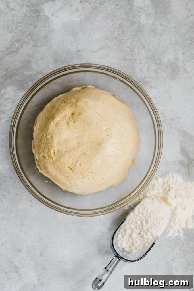 Soft Parmesan Dinner Roll dough resting in a glass bowl, beside a scoop of flour, demonstrating the initial stage of bread making.