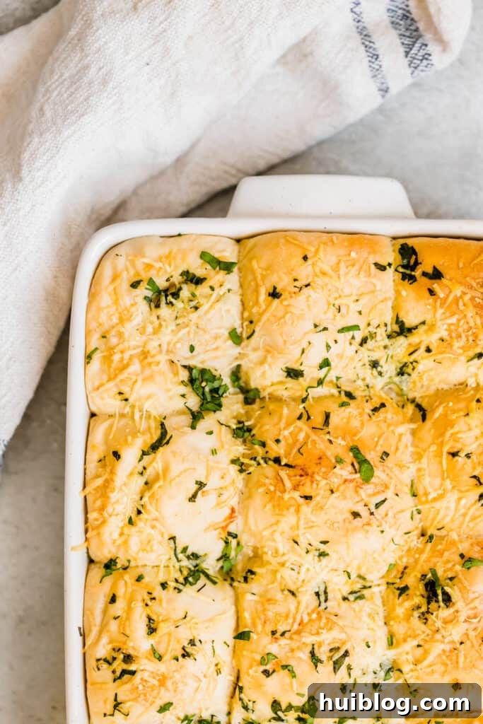 A pan overflowing with freshly baked dinner rolls, their tops glistening with melted Parmesan and flecks of green parsley.