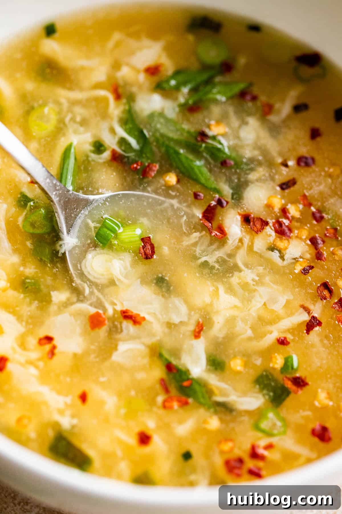 Close-up side view of egg drop soup in a white bowl, garnished with chopped green onions and a sprinkle of red pepper flakes.