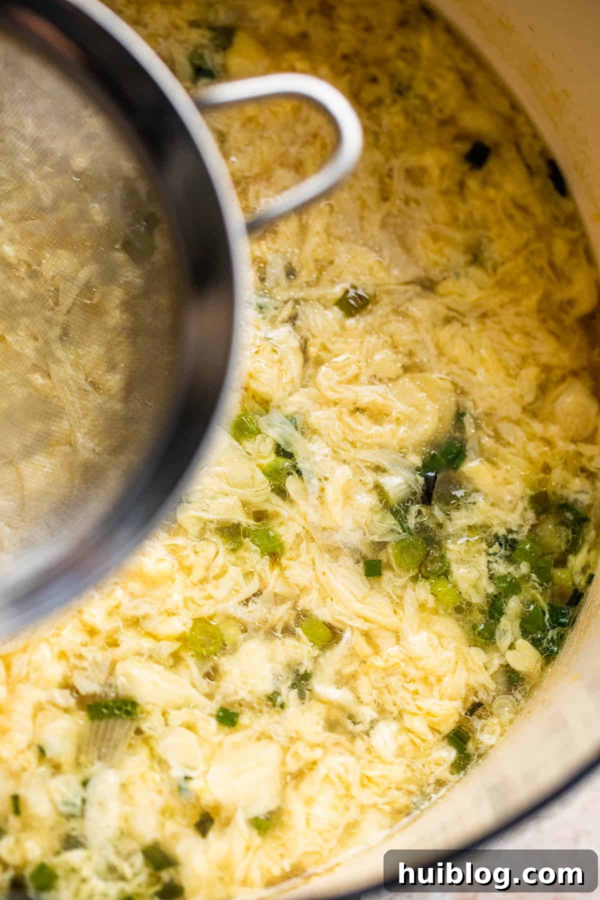 A hand gently drizzling beaten egg through a wire mesh sieve into a simmering pot of broth to create delicate egg ribbons for egg drop soup.
