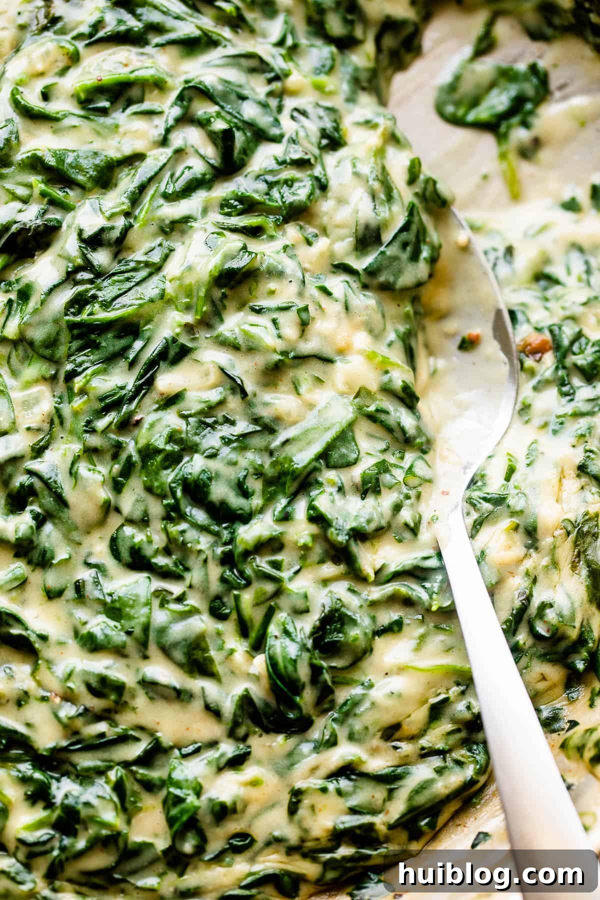 A large spoon stirring rich, thick creamed spinach in a stainless steel skillet, showcasing its creamy texture.