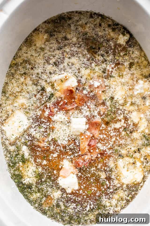 All the delicious ingredients - chicken broth, cream cheese cubes, bacon, chicken breasts, and seasonings - ready in the slow cooker before cooking.