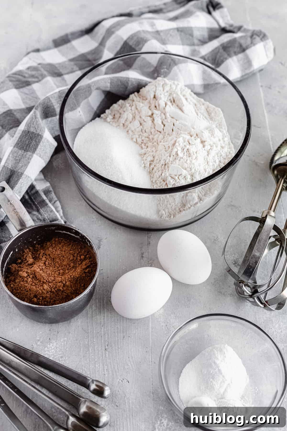 Arrangement of cocoa powder, eggs, baking soda, baking powder, and other cake ingredients on a gray countertop